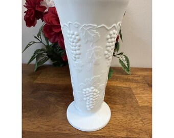 May include: White ceramic vase with a flared top and a round base. The vase features an embossed grape and leaf design. The vase is displayed on a wooden surface with red and white flowers in the background.