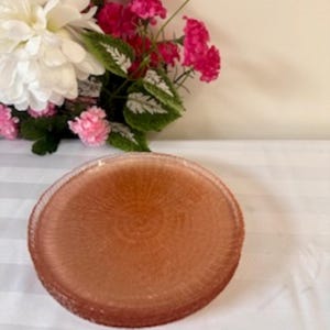 Puede incluir: Una pila de platos redondos de vidrio color ámbar. Los platos tienen una superficie texturizada y un borde decorativo. Los platos están sobre una superficie blanca con un arreglo floral en el fondo.