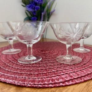 Puede incluir: Cuatro copas de postre de vidrio transparente con tallo y un patrón floral sobre un mantel tejido rosa.