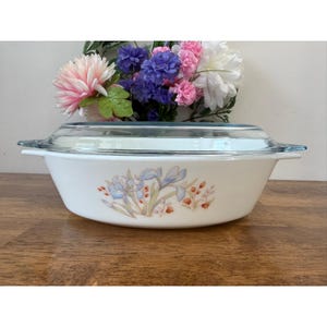 May include: White oval casserole dish with a clear glass lid. The dish features a floral design with blue and orange flowers. The dish is sitting on a wooden surface.