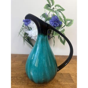 May include: A teal and black ceramic jug with a curved handle. The jug has a teardrop shape and a glossy finish. The handle and top are black, while the body transitions from teal to a darker shade. Decorative flowers are in the background.