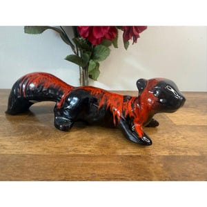 May include: A ceramic figurine of a long-bodied animal, possibly a ferret or weasel, featuring a glossy black base glaze with vibrant orange-red drip patterns across its back, tail, and head, captured in a horizontal, low-profile stance.