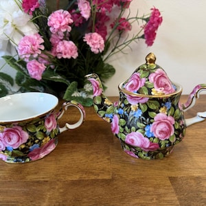 Könnte beinhalten: Ein florales Teeservice mit Teekanne und Tasse. Das Set hat einen schwarzen Hintergrund mit rosa Rosen, blauen Akzenten und goldenen Verzierungen. Die Tasse und die Teekanne stehen auf einer Holzoberfläche.