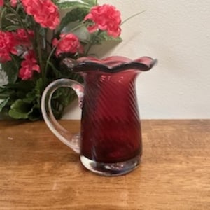 May include: A small, ruby red glass pitcher with a clear handle and a ruffled rim. The pitcher has a spiral design and sits on a wooden surface. Decorative red flowers are in the background.