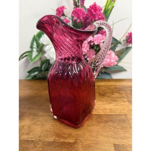 May include: A ruby red glass pitcher with a textured, swirled design. The handle is clear and twisted. The pitcher has a square base and a flared spout. The background includes pink flowers and greenery.