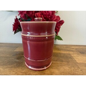 Può includere: Un barattolo in ceramica bordeaux con coperchio abbinato. Il barattolo ha una forma cilindrica con la parte superiore e inferiore leggermente svasate. Una fascia beige chiaro circonda il centro. Lo sfondo presenta fiori rossi.
