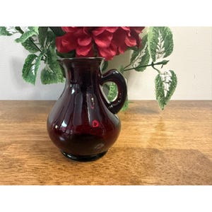 Peut inclure: Une petite cruche en verre rouge foncé avec une anse. La cruche a un corps arrondi et un bord légèrement évasé. Elle repose sur une surface en bois, avec des fleurs rouges et des feuilles vertes en arrière-plan. La cruche est probablement à usage décoratif.