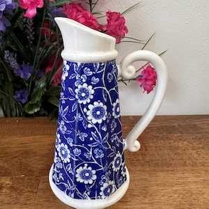 May include: A blue and white floral patterned jug with a white handle and rim. The jug has a flared spout and a decorative floral design. The jug is a decorative item.