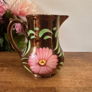 May include: A brown ceramic pitcher with a gold handle and spout. The pitcher is decorated with a large pink flower and green leaf accents. The pitcher is sitting on a wooden surface.