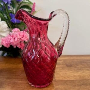 May include: A small, ruby red glass jug with a clear glass handle. The jug has a diamond pattern and is ideal for serving juice or water.