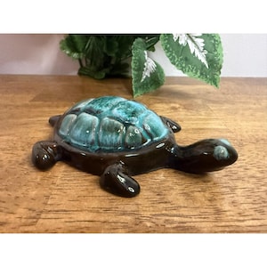 Może przedstawiać: Ceramiczna figurka żółwia z błyszczącym wykończeniem. Skorupa żółwia to mieszanka turkusu i zieleni, a korpus i głowa są ciemnobrązowe. Figurka jest wystawiona na drewnianej powierzchni.