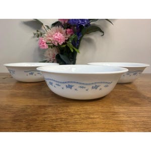 Peut inclure: Trois bols en céramique blanche avec un motif floral bleu sur le bord. Les bols sont disposés sur une surface en bois, avec un vase de fleurs en arrière-plan.
