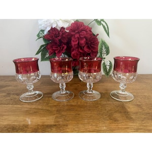 Può includere: Quattro calici da vino vintage con coppe rosso rubino e steli trasparenti a motivi. I bordi sono accentuati con oro. I calici sono disposti su una superficie di legno, con una composizione floreale sullo sfondo.