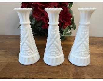 Puede incluir: Tres jarrones blancos de vidrio lechoso con un diseño geométrico texturizado. Los jarrones tienen una parte superior acampanada y un patrón detallado. Los jarrones están dispuestos sobre una superficie de madera con un fondo de flores rojas.