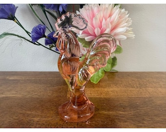 May include: A translucent pink glass figurine shaped like a rooster, featuring a detailed comb, wattle, and curved tail feathers. The glass sculpture stands on a circular base and has a smooth, reflective surface throughout its entire form.