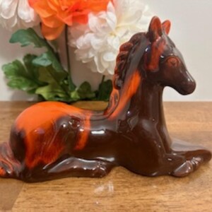 Puede incluir: Una figurita de caballo de cerámica en un esmalte marrón y naranja. El caballo está acostado con la cabeza girada hacia un lado.