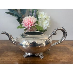 Peut inclure: Une théière argentée avec une anse et un bec courbés, reposant sur trois pieds ornés. La théière est très réfléchissante, avec une ouverture sombre sur le dessus. Le fond présente une surface en bois et une composition florale avec des fleurs roses et blanches.