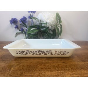Puede incluir: Fuente rectangular de color crema con un diseño floral marrón alrededor del borde. La fuente está vacía y descansa sobre una superficie de madera. Flores decorativas en el fondo.
