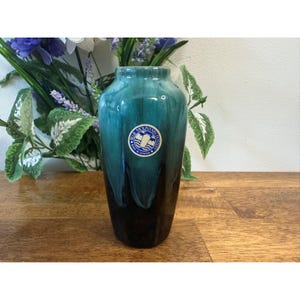 Puede incluir: Un jarrón alto de cerámica vidriada en color verde azulado y negro. El jarrón tiene una etiqueta que dice "Blue Mountain Pottery, Made in Canada". El jarrón se exhibe sobre una superficie de madera, con vegetación y flores en el fondo.