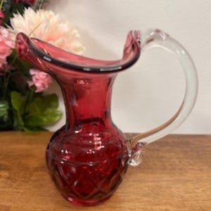 May include: A ruby red glass jug with a clear glass handle. The jug has a flared rim and a diamond pattern on the lower portion. The handle is arched and attached to the side of the jug. The jug is likely used for serving beverages.