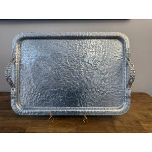 May include: A rectangular, hammered aluminium serving tray with decorative handles. The tray features a textured surface and an embossed floral design in the centre. The tray is displayed on a wooden surface.