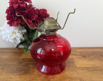 May include: A vintage red glass oil lamp with a brass burner and handle. The lamp has a round body and a pedestal base. The background includes red and white flowers on a wooden surface.