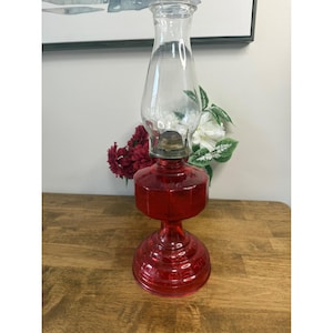 May include: A vintage red glass oil lamp with a clear glass chimney. The lamp has a round base and a hexagonal body. The lamp is approximately 30 cm tall. The lamp is sitting on a wooden surface.