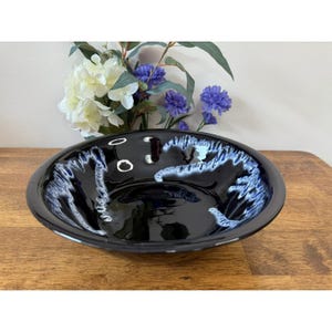 Puede incluir: Un cuenco de cerámica negro brillante con un diseño único. El interior presenta un llamativo patrón de vetas blancas y azules sobre un fondo oscuro. El cuenco, de forma ovalada, está sobre una superficie de madera.