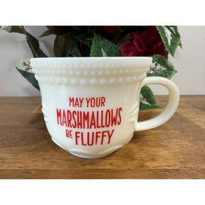 Könnte beinhalten: Cremefarbene Keramiktasse mit runder Form und Henkel. Die Tasse trägt den Text "MAY YOUR MARSHMALLOWS BE FLUFFY" in roter Schrift. Die Tasse hat ein Perlendesign am Rand und dekorative Akzente an den Seiten.