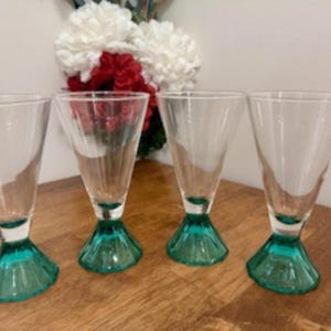 May include: Four clear glass drinking glasses with teal green bases. The glasses are arranged in a row on a wooden surface.