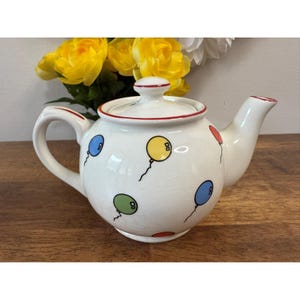 May include: White ceramic teapot with a lid, handle, and spout. The teapot is decorated with colourful balloon designs in blue, yellow, green, and red. The rim and lid are trimmed with red. A charming addition to any kitchen.