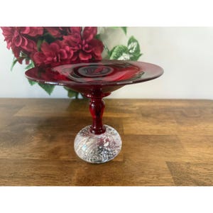 Peut inclure: Un coupe à pied en verre rouge rubis avec un dessus rond et plat et une base bulbeuse et transparente avec des paillettes irisées. Le bol a une courte tige et est exposé sur une surface en bois. Le bol est probablement utilisé pour servir ou exposer.