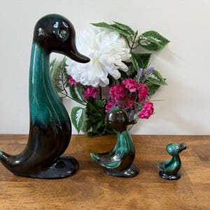 Könnte beinhalten: Drei Keramik-Entenfiguren in verschiedenen Größen. Die Enten haben eine glänzende Oberfläche mit einem Farbverlauf von Dunkelbraun zu Türkis. Eine große Ente steht hoch, während zwei kleinere Enten in der Nähe positioniert sind. Im Hintergrund befindet sich ein Blumenarrangement.