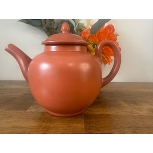 May include: A terracotta-colored teapot with a matching lid and handle. The teapot is round with a curved spout and a rounded handle. The background features a wooden surface and colorful flowers.