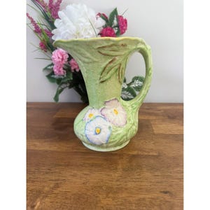 May include: A light green ceramic pitcher with a handle and a floral design. The pitcher features a bulbous base and a flared top. The floral design includes white, blue, and purple flowers with yellow centers. The pitcher is approximately 7 inches tall.