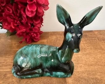 May include: A ceramic deer figurine in shades of green and black, with a glossy finish. The deer is in a resting position with its ears perked up. Red flowers are in the background.
