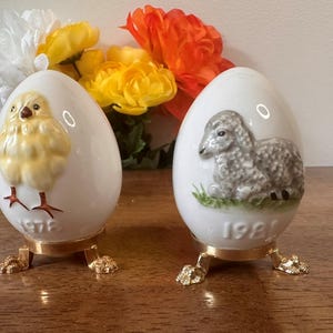 Puede incluir: Dos adornos de huevo de Pascua de cerámica blanca. Uno presenta un pollito amarillo, el otro un cordero gris. Ambos están sobre soportes dorados y tienen los años 1978 y 1981 impresos.