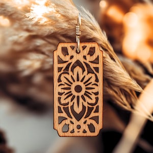 May include: A wooden earring with a delicate floral design. The earring is a rectangular shape with a cutout design that resembles a flower. The earring is made of light brown wood and has a gold-colored hook.