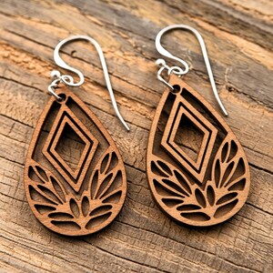 May include: A pair of wooden teardrop earrings with a geometric design. The earrings feature a diamond shape in the center and a floral-like pattern at the bottom. The earrings hang from silver-coloured ear wires.