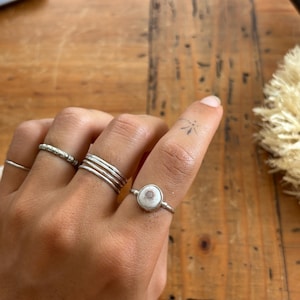 May include: A set of silver-toned rings worn on fingers. One ring features a small, circular, white and brown shell-like inlay. The other rings include a thin plain band, a textured band, and a stack of three thin, smooth, silver-toned bands.