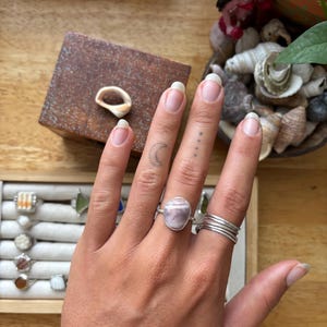 May include: A hand wearing several rings, including a silver ring with a large, oval, light purple stone. Other rings are simple silver bands. A wooden box with a seashell is in the background, along with a tray of rings.