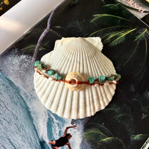 May include: A double-strand bracelet featuring raw turquoise nuggets and small white pearls strung on a thin orange cord, centered with a single circular white seashell bead, resting on a large, ribbed white scallop shell.