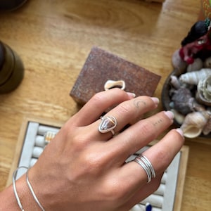 May include: A hand wearing a silver ring with a teardrop-shaped, brown and white stone. The hand also has several silver rings and a silver bracelet. The background includes a wooden box and a tray with jewelry.