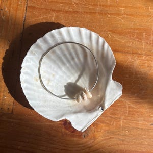 May include: A silver bangle bracelet with three small, pale seashells rests inside a large, white seashell. The seashell is on a wooden surface. The bracelet and shells are a decorative item.