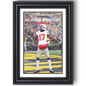 May include: Framed print of The Columbus Dispatch newspaper cover, featuring a football player in a white and red uniform with the number 17. The headline reads "12-OVER MICHIGAN". The image is set against a stadium backdrop.