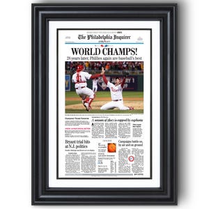 May include: Framed newspaper front page with the headline "WORLD CHAMPS!" celebrating a baseball victory. The image features a black frame and a white mat, with action shots of the game and additional news articles.