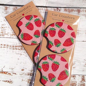 May include: Two sets of car coasters with a pink background and a strawberry pattern. The coasters are packaged in clear plastic and have a brown paper backing with the text "Car Coasters Handmade with love".