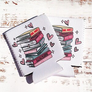 May include: Two white beverage insulators with pink hearts and stars. The insulators feature a stack of books with the text "Soul Mates", "Forbidden Love", "Fated Mates", "Love, Actually", "Slow Burn", and "Make Me Burn Like You" on the covers.
