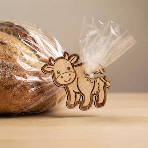 May include: A light brown wooden bread clip shaped like a cow with dark brown laser-etched outlines and facial features. The clip is attached to a clear plastic bag containing a round loaf of crusty bread, securing the gathered plastic top.