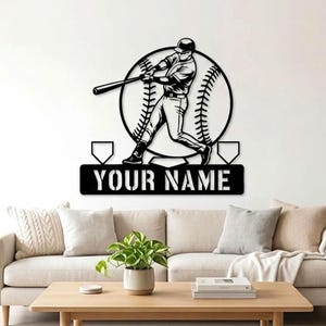 May include: A black metal wall art piece featuring a silhouette of a baseball player swinging a bat in front of a large baseball graphic. Below the player, a rectangular plaque displays the text YOUR NAME flanked by two home plate shapes.